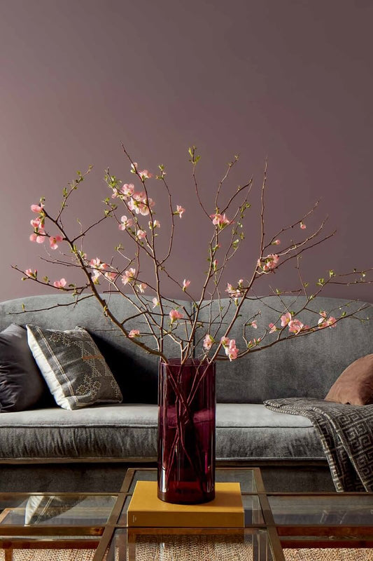 A purple-painted living room features a gray couch, gold side tables, and a glass-topped gold coffee table topped with a red vase filled with pink flowers.